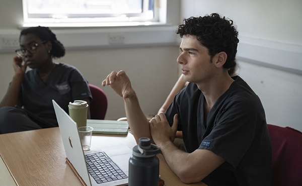 Image of a white man with dark hair wearing scrubs, sitting at a desk in front of a computer